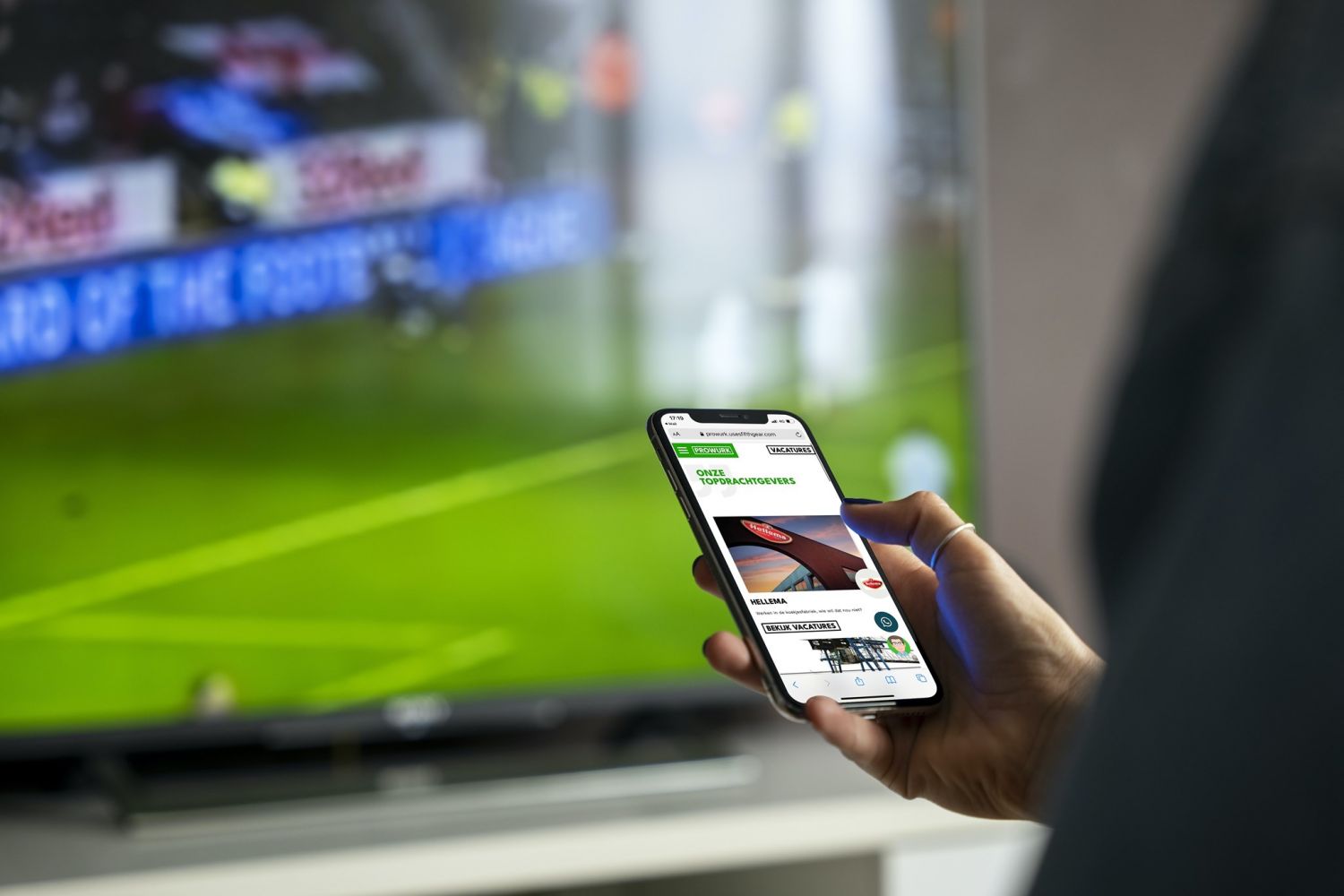De homepage van ProWurk op een mobiel met een voetbalwedstrijd op een tv op de achtergrond